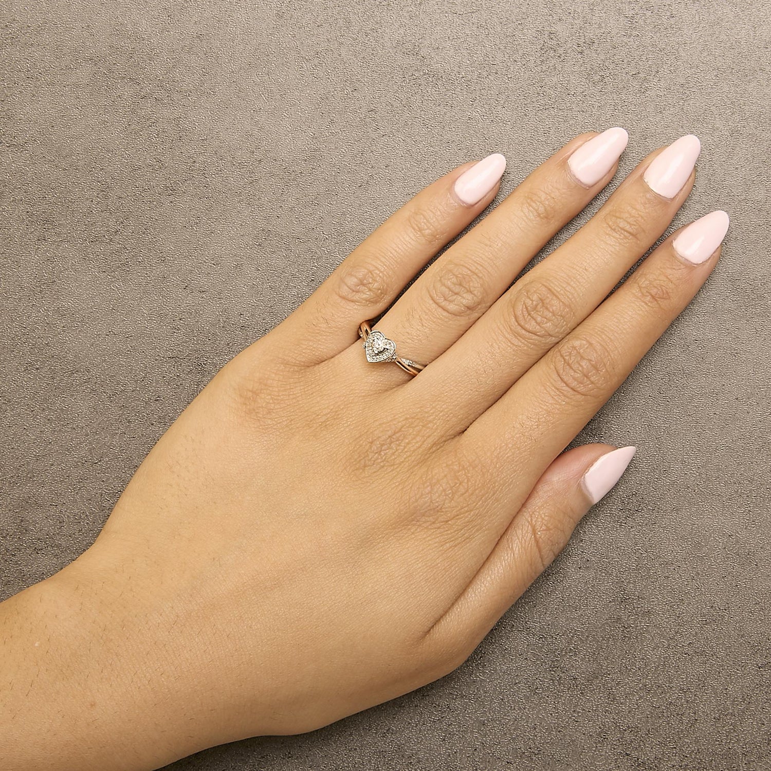 Hand with pink nails wearing a heart-shaped diamond ring on a textured beige surface.