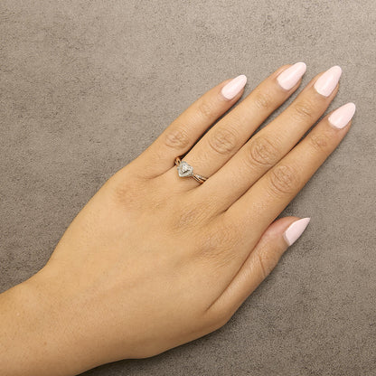 Hand with pink nails wearing a heart-shaped diamond ring on a textured beige surface.