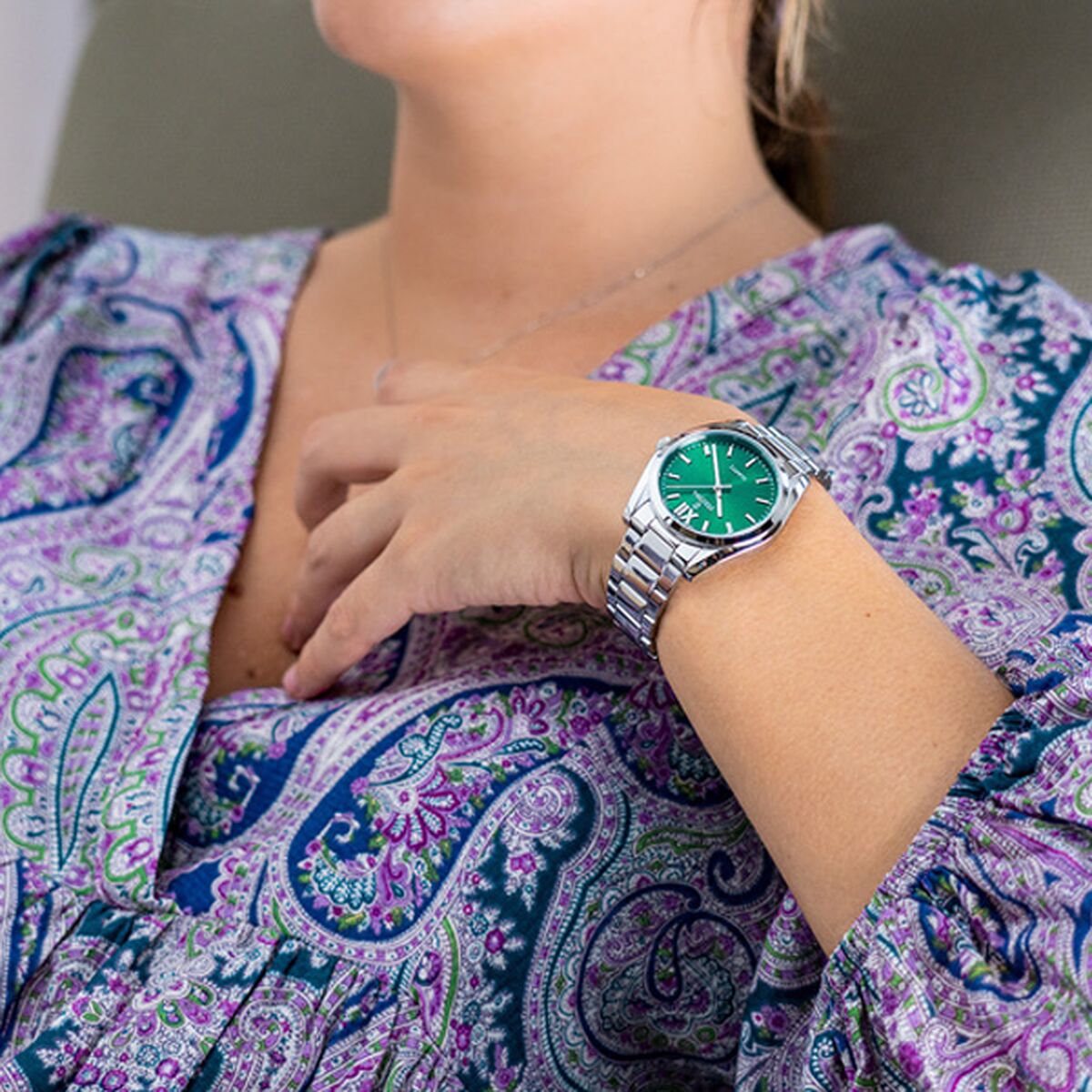 Woman wearing a silver watch with a green face, purple paisley patterned blouse in background.