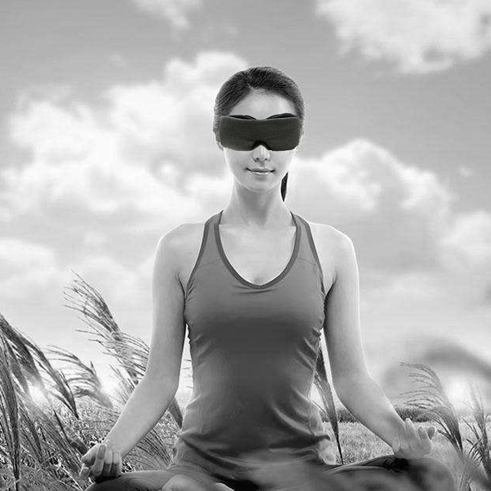 Woman meditating in a field, wearing a sleeveless top and eye mask, with a cloudy sky backdrop.