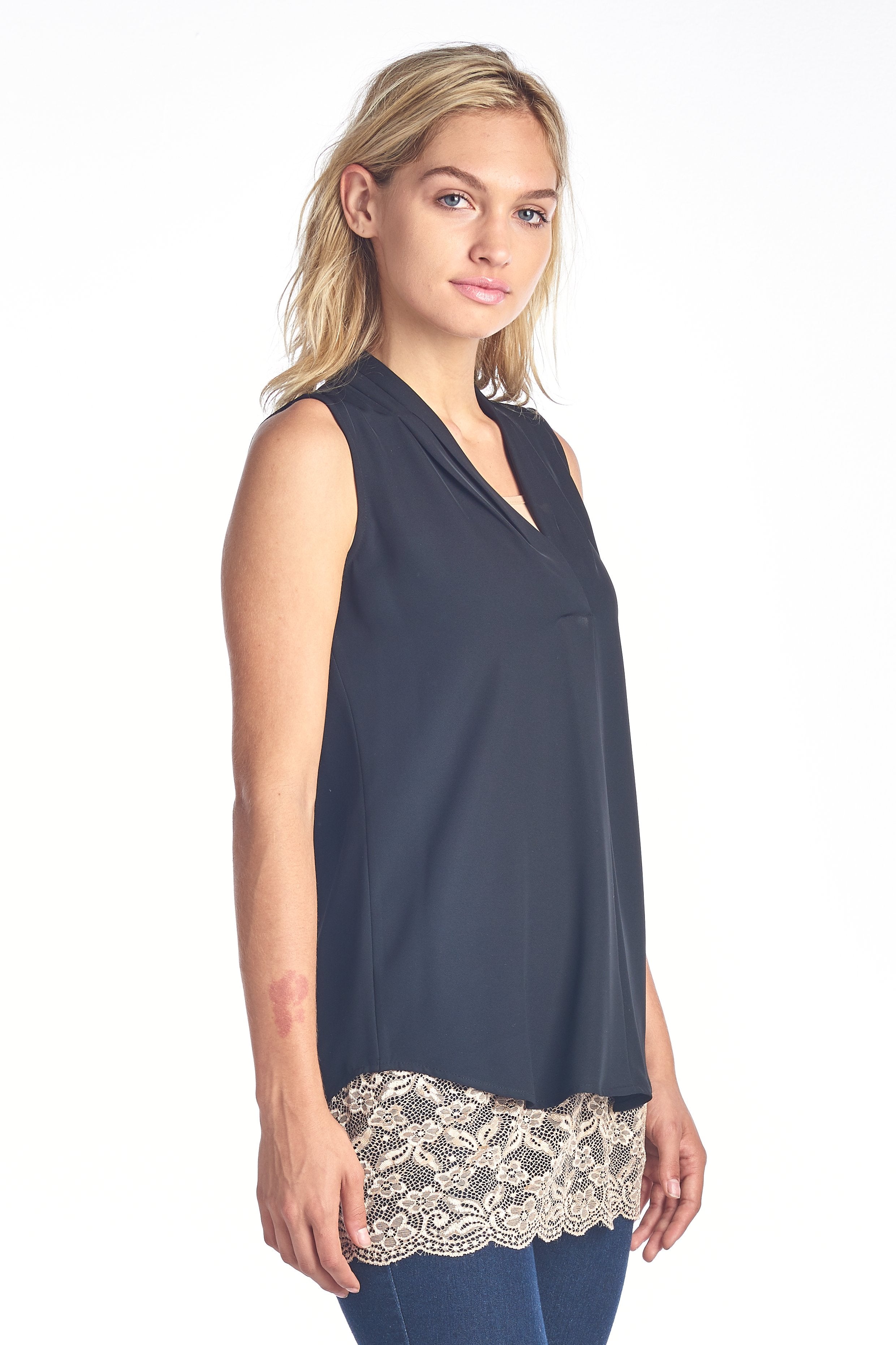 Woman in black sleeveless top with lace trim, wearing blue jeans, standing against a white background.