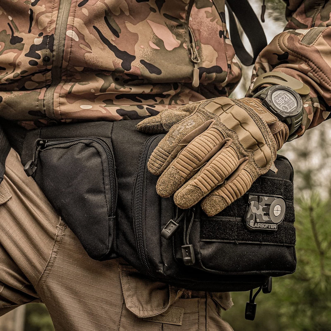 Person in camo jacket with brown gloves, black tactical pouch, and wrist watch outdoors.