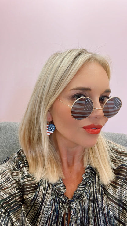Blonde woman in striped top, wearing American flag sunglasses and earrings, pink background.
