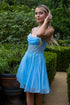 Woman in a blue, embellished dress stands in a lush garden, touching her hair.