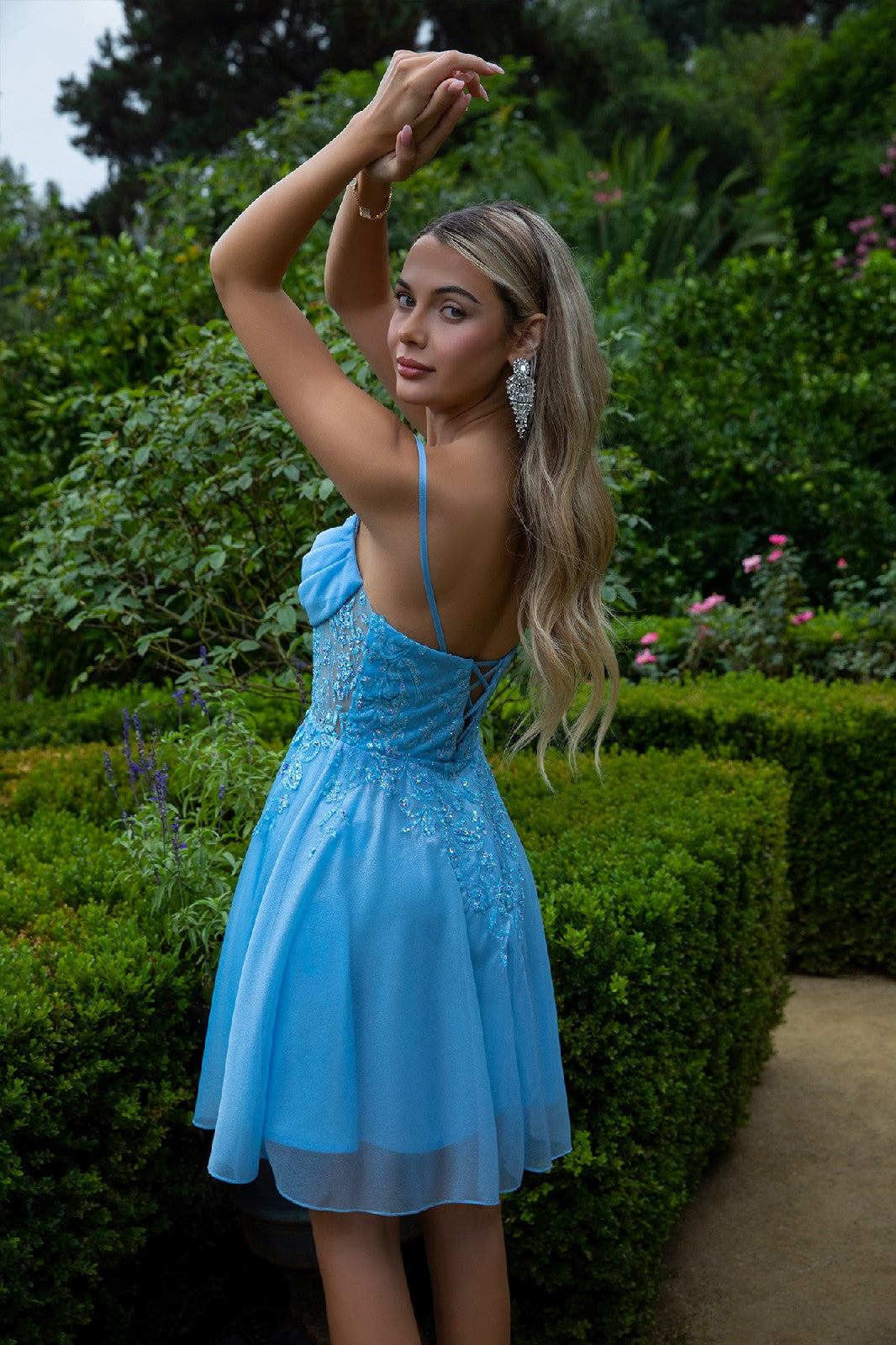 Woman in blue dress poses with arms raised, surrounded by lush green garden foliage.