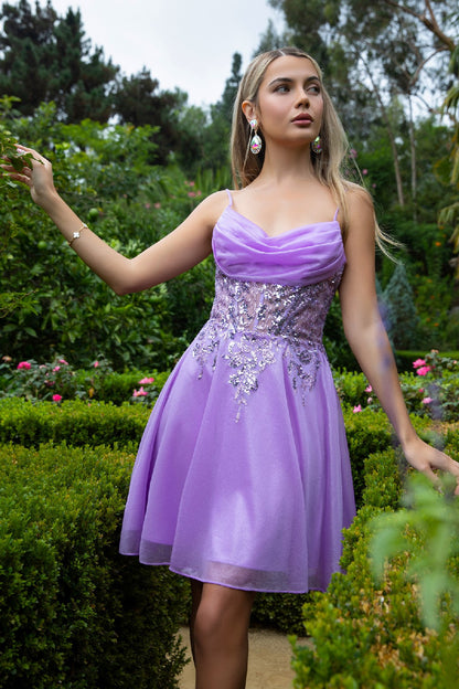 Woman in purple floral dress standing in garden with green hedges and lush foliage.