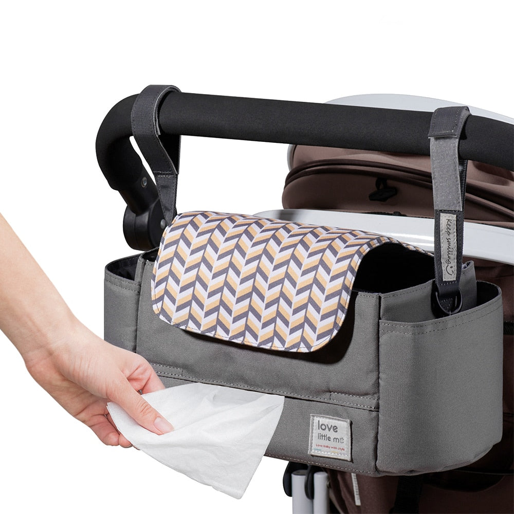 Gray stroller caddy with chevron flap, attached to handle. Hand pulls wipe from the dispenser.