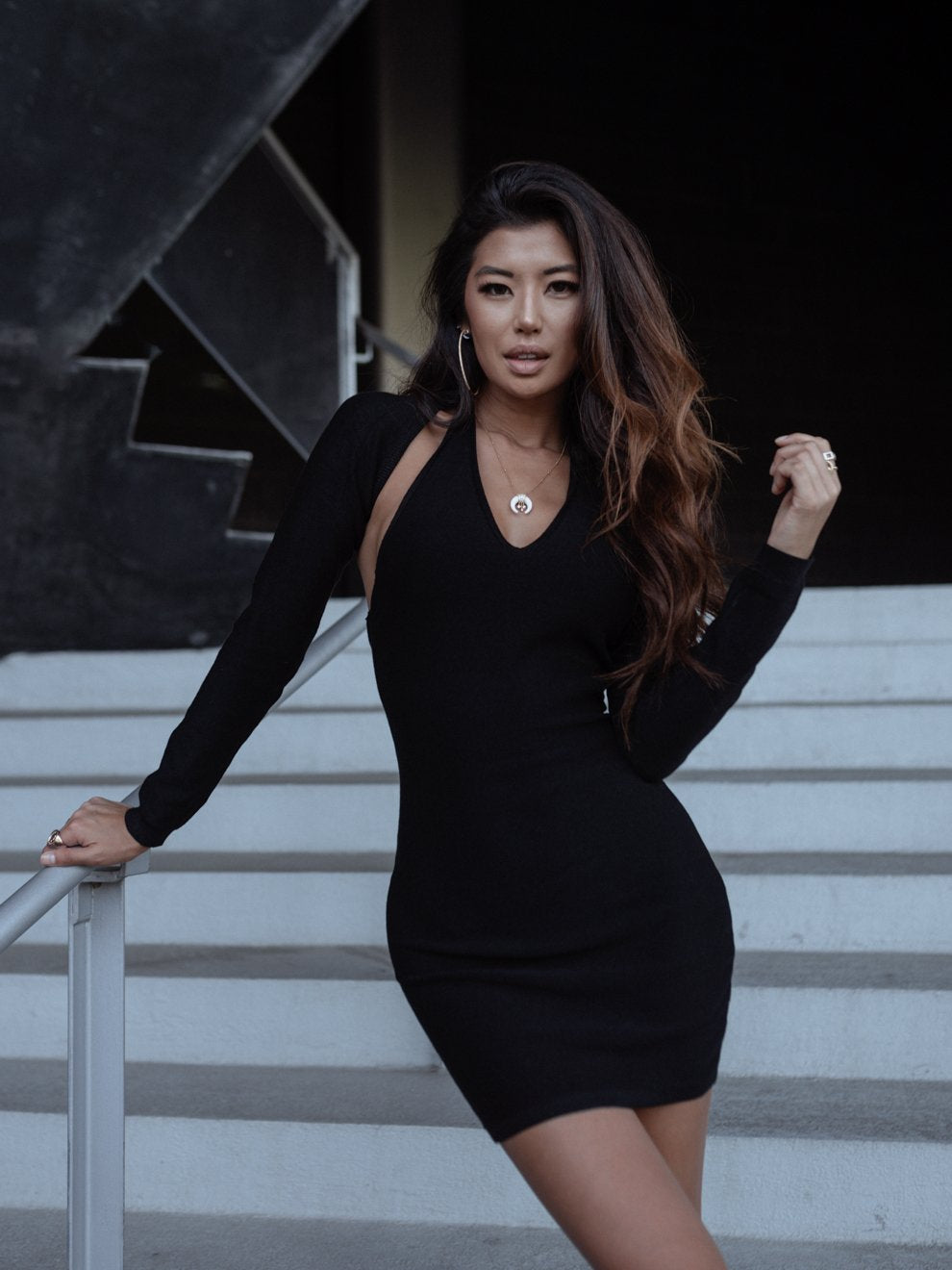 Woman in a black dress standing on stairs, hand on railing, long hair cascading over shoulder.
