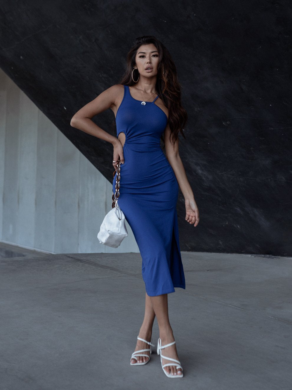 Woman in a blue dress with hoop earrings, holding a white bag, against a dark background.