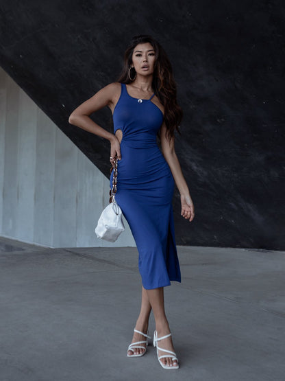 Woman in a blue dress with hoop earrings, holding a white bag, against a dark background.