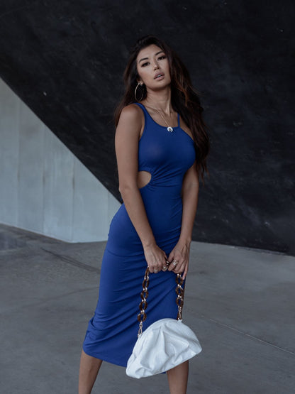 Woman in a blue dress holding a white purse with chain handles against a dark background.