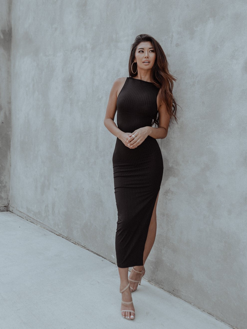Woman in a black dress with side slit, leaning against a gray textured wall, wearing hoop earrings.