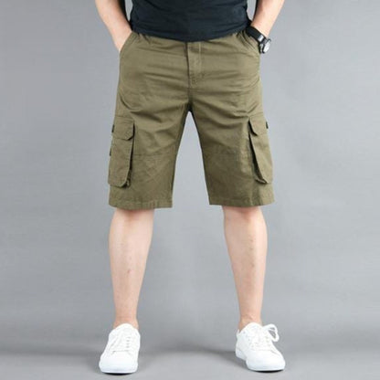 Man in olive cargo shorts, black shirt, and white sneakers standing against a gray background.