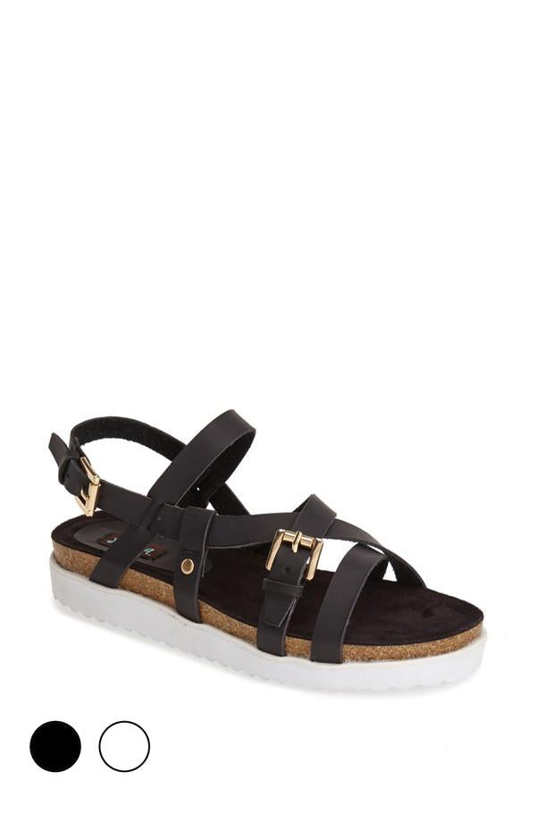 Black sandal with gold buckles and white sole on a plain background.