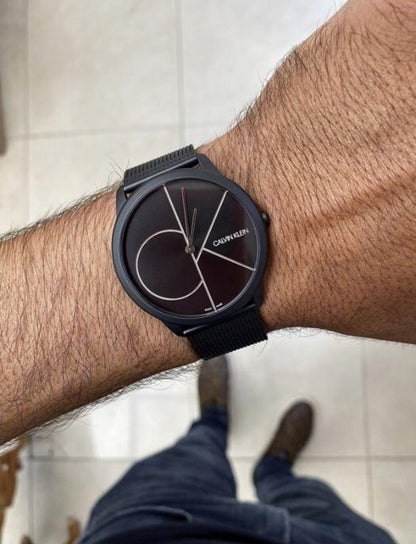 Black wristwatch with a minimalist face and silver lines on a wrist, wearing dark jeans and boots.