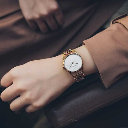 Wrist with gold watch and white face, wearing a brown sleeve, resting on dark surface.
