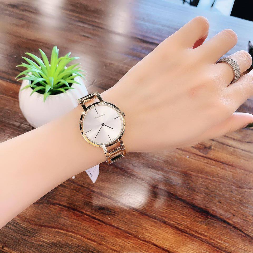 Wrist with a gold watch and silver dial on a wooden table; potted plant in the background.
