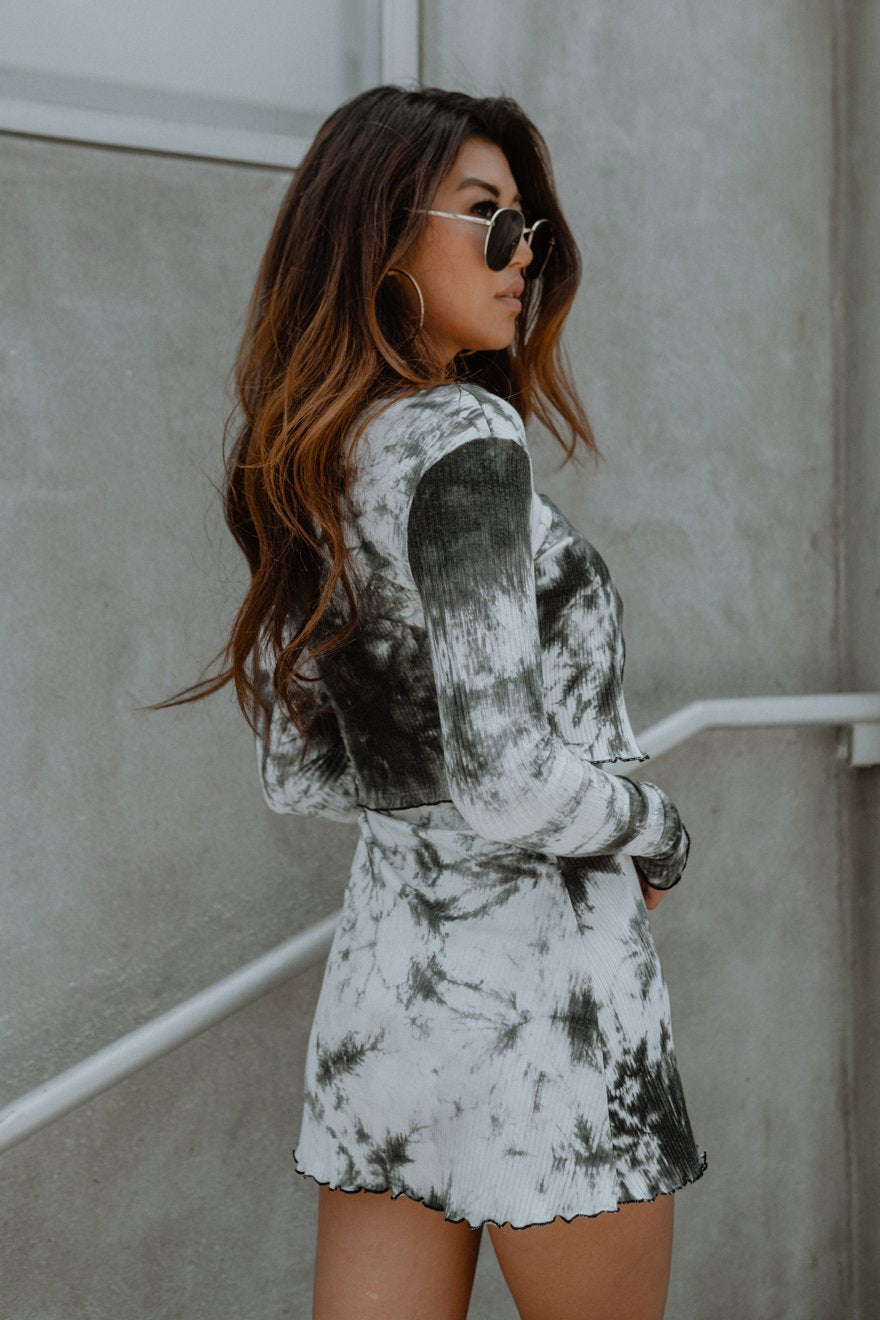 Woman with wavy hair in tie-dye dress, wearing sunglasses, against a concrete wall.