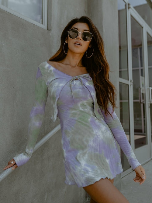 Woman in a pastel tie-dye dress with sunglasses, leaning against a concrete wall near glass doors.