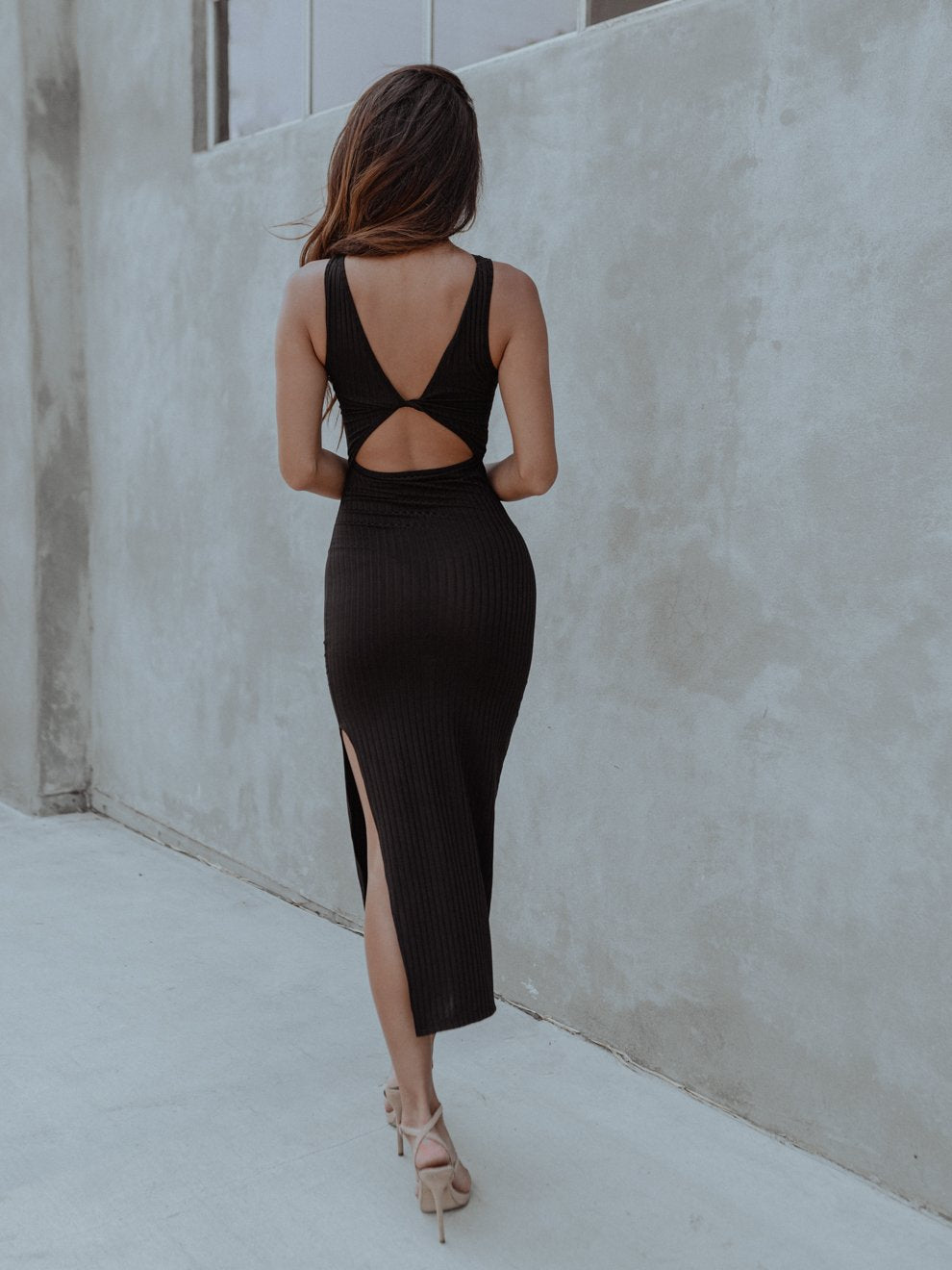 Woman in a black dress with a back cutout, walking on a light concrete floor beside a gray wall.