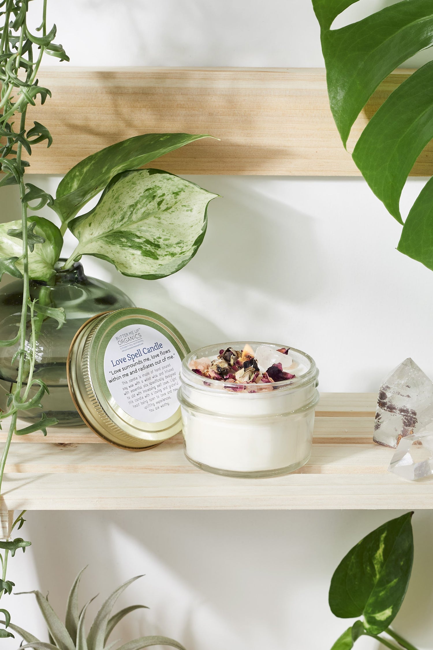 Jar candle with rose petals on a wooden shelf, surrounded by green plants and crystals.
