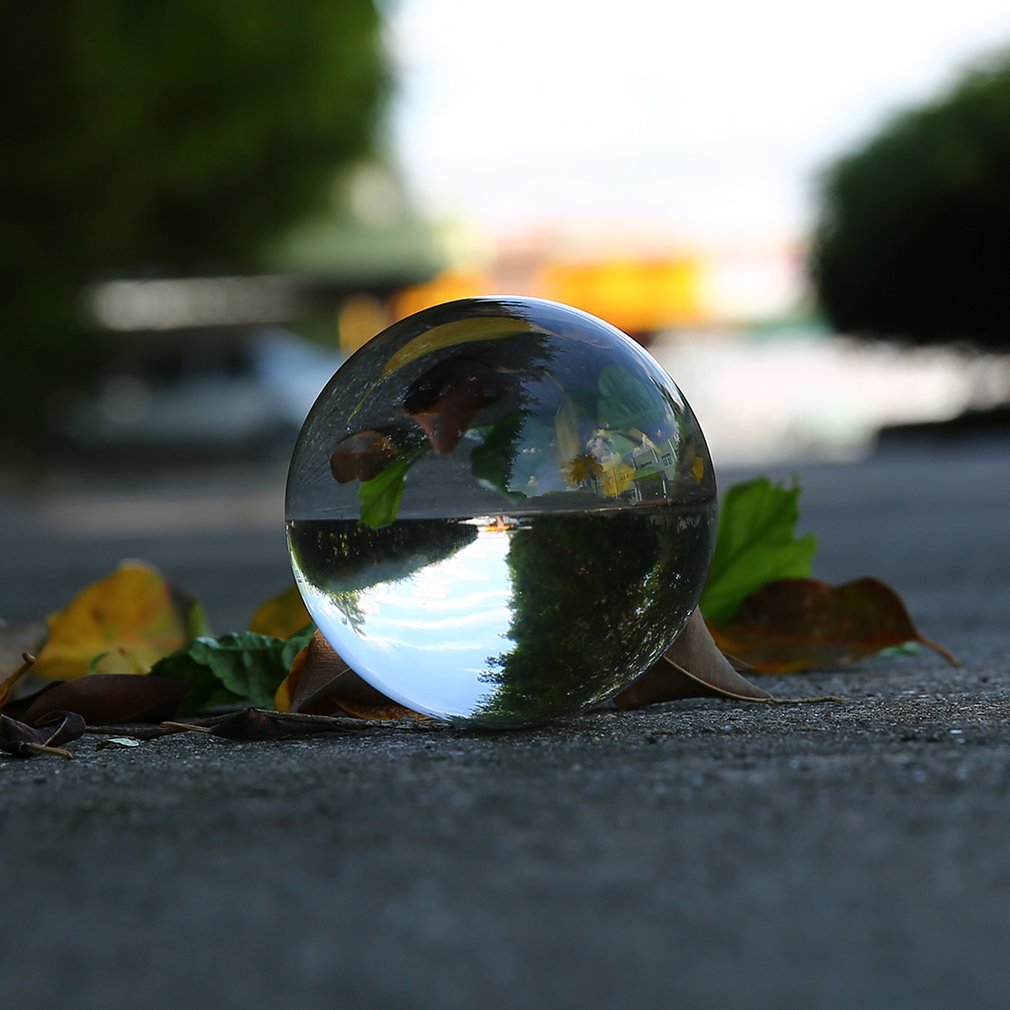 Glass sphere on pavement, reflecting trees and sky; leaves surround it, creating a vibrant scene.
