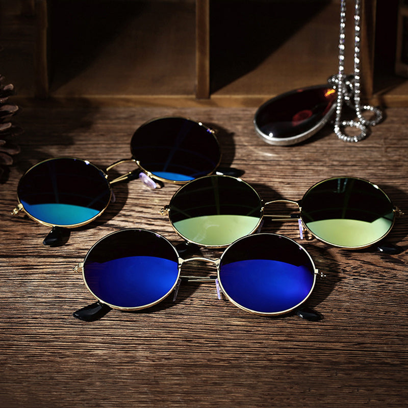 Round sunglasses with blue and green lenses on a wooden table, beside a chain and pendant.