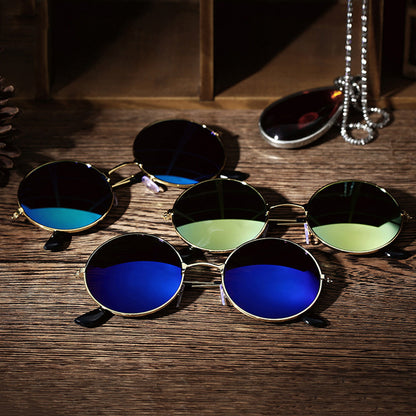 Round sunglasses with blue and green lenses on a wooden table, beside a chain and pendant.