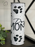 White tumbler with black paw prints and "Dog Mom" text. Green leaves in the background.