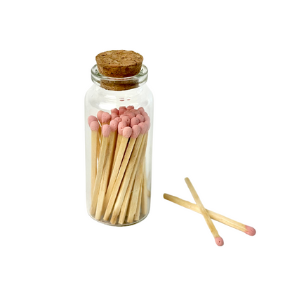 Glass jar with cork filled with matches, pink tips; two matches lie beside it.