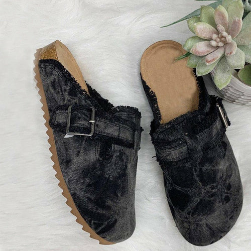 Black slip-on clogs with frayed edges and a buckle next to a succulent plant on white background.