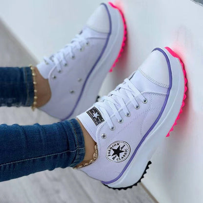 White sneakers with purple trim and neon pink soles, worn with blue jeans against a white wall.