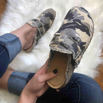 Camouflage clogs with frayed edges, worn by a person in jeans on a white plush rug.