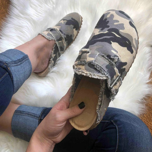 Camouflage slip-on shoes with a frayed edge on a white fluffy rug, worn with blue jeans.