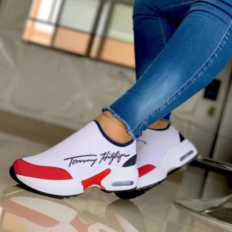 White sneakers with red and black accents worn with blue jeans, reflected on a shiny floor.