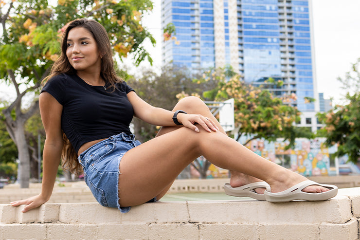 Woman in a black top and denim shorts sits on bricks, with tall buildings and trees in the background.