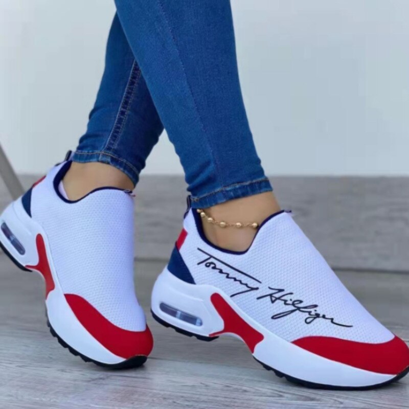 White slip-on sneakers with red and navy accents, worn with blue jeans on a light wooden floor.