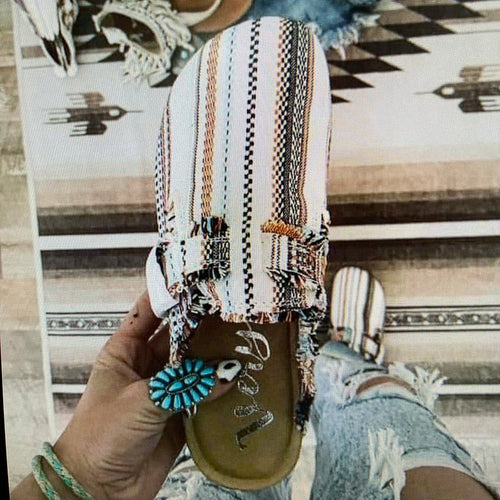 Hand holding a striped, multicolored shoe with a turquoise bird charm; ripped jeans visible.
