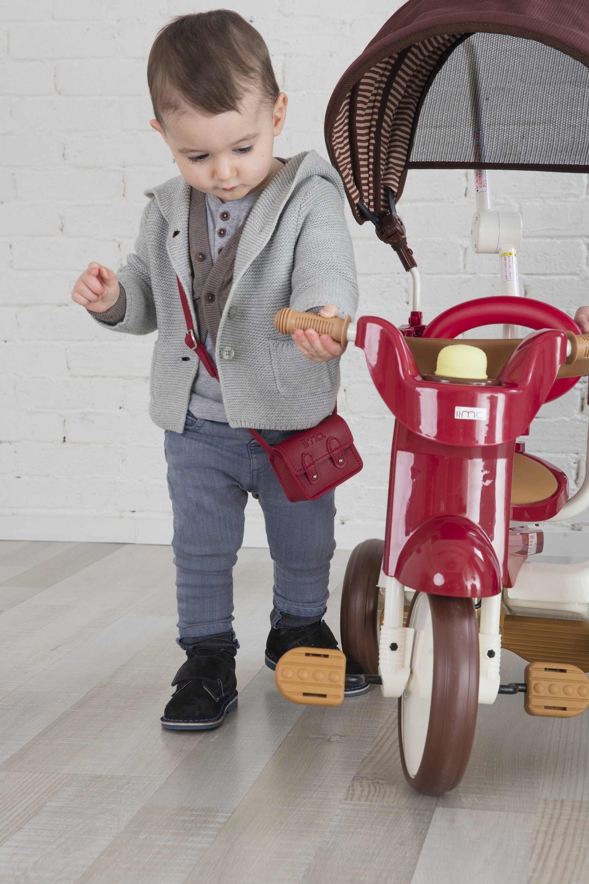 Child in grey sweater with red bag holding a red tricycle handle indoors.