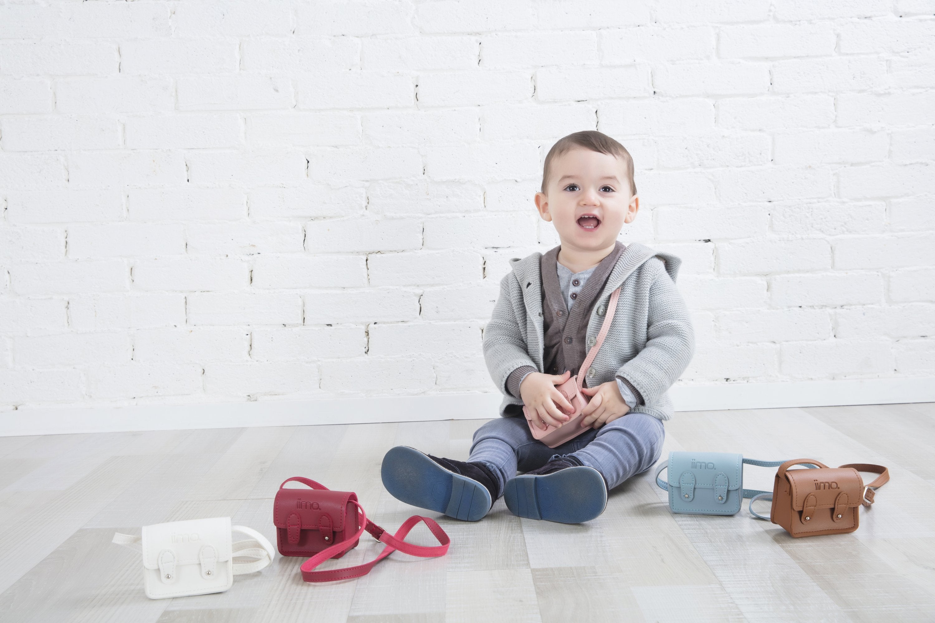 Toddler in gray sweater sits on the floor with colorful mini bags against a white brick wall.