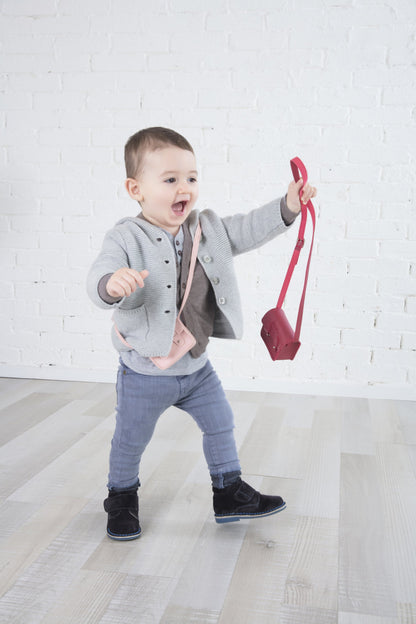 Toddler in grey cardigan and jeans, holding a red purse, smiling in a bright room with white wall.