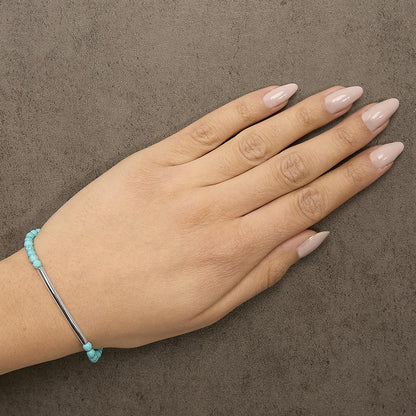 Hand with nude nails and a turquoise bead bracelet on a textured gray surface.