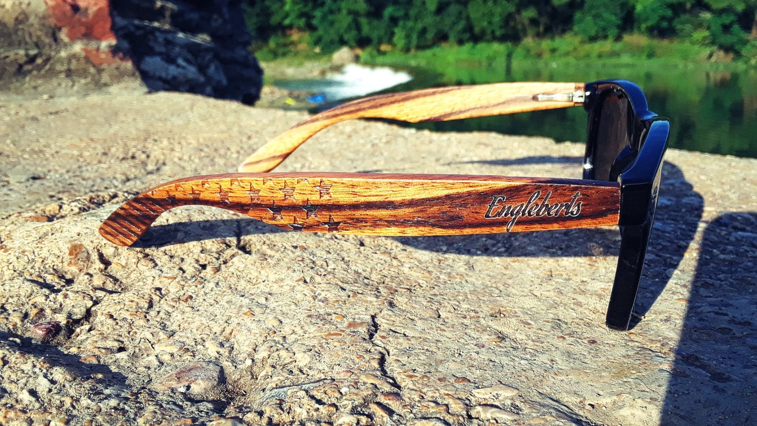 Wood-grain sunglasses on textured stone, forest and water in the background.