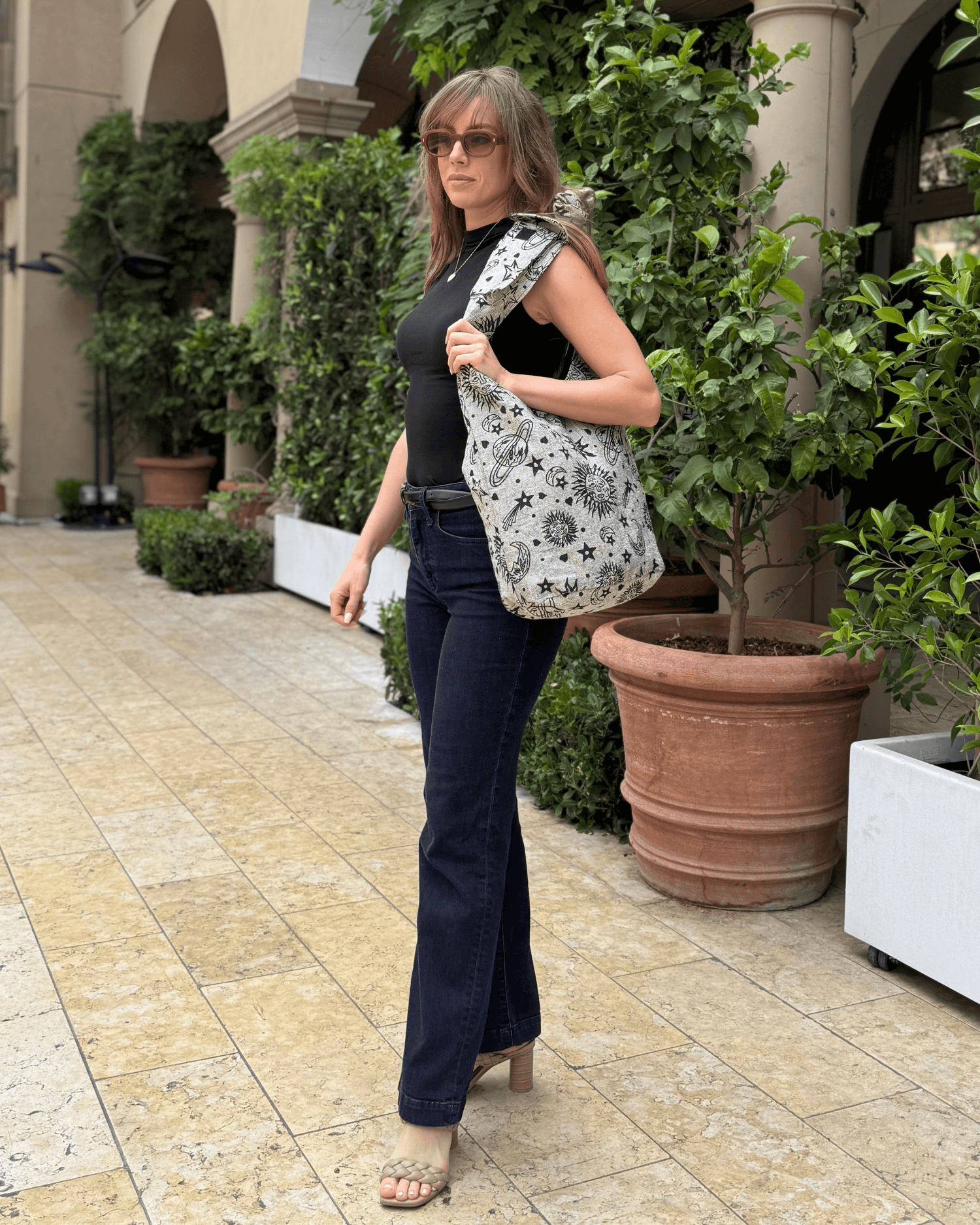 Woman in black top and jeans, carrying a star-patterned bag, walking by potted plants.