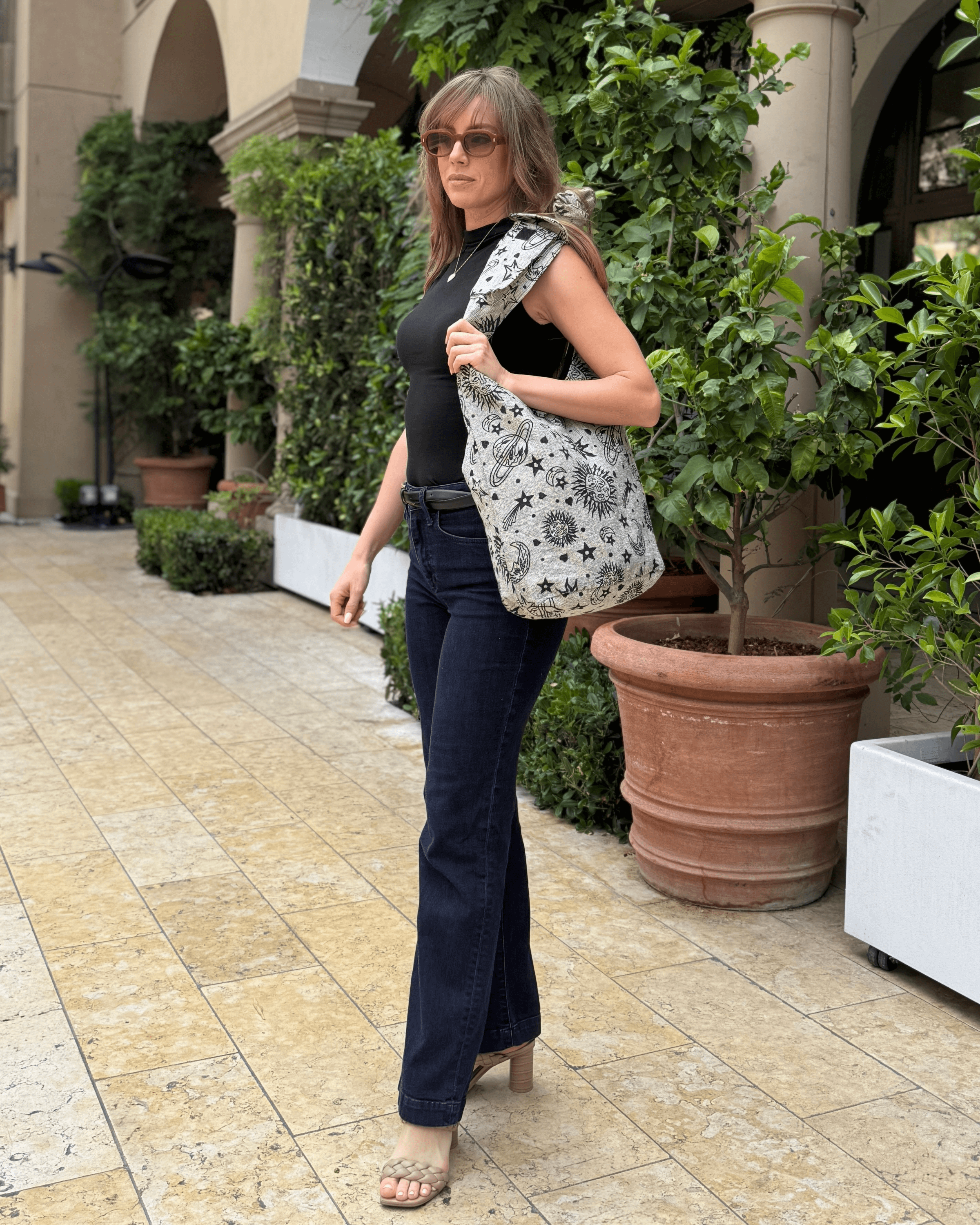 Woman in black top and jeans, carrying a star-patterned bag, walking by potted plants.