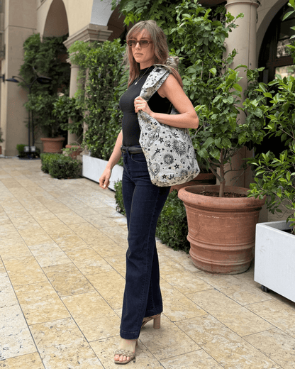 Woman in black top and jeans, carrying a star-patterned bag, walking by potted plants.