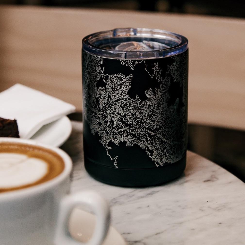 Black tumbler with intricate white line pattern on table, near cappuccino and brownie.