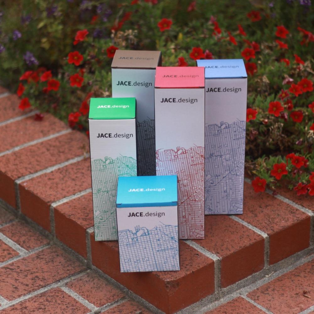 Colorful JACE.design boxes with city map patterns on brick steps, surrounded by red flowers.