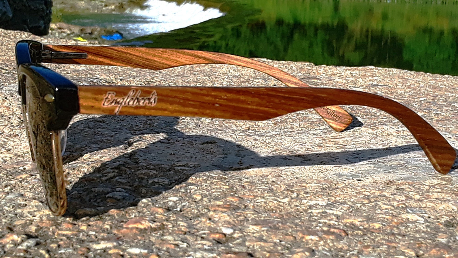 Wooden sunglasses with black frames rest on textured stone, greenery and water in the background.