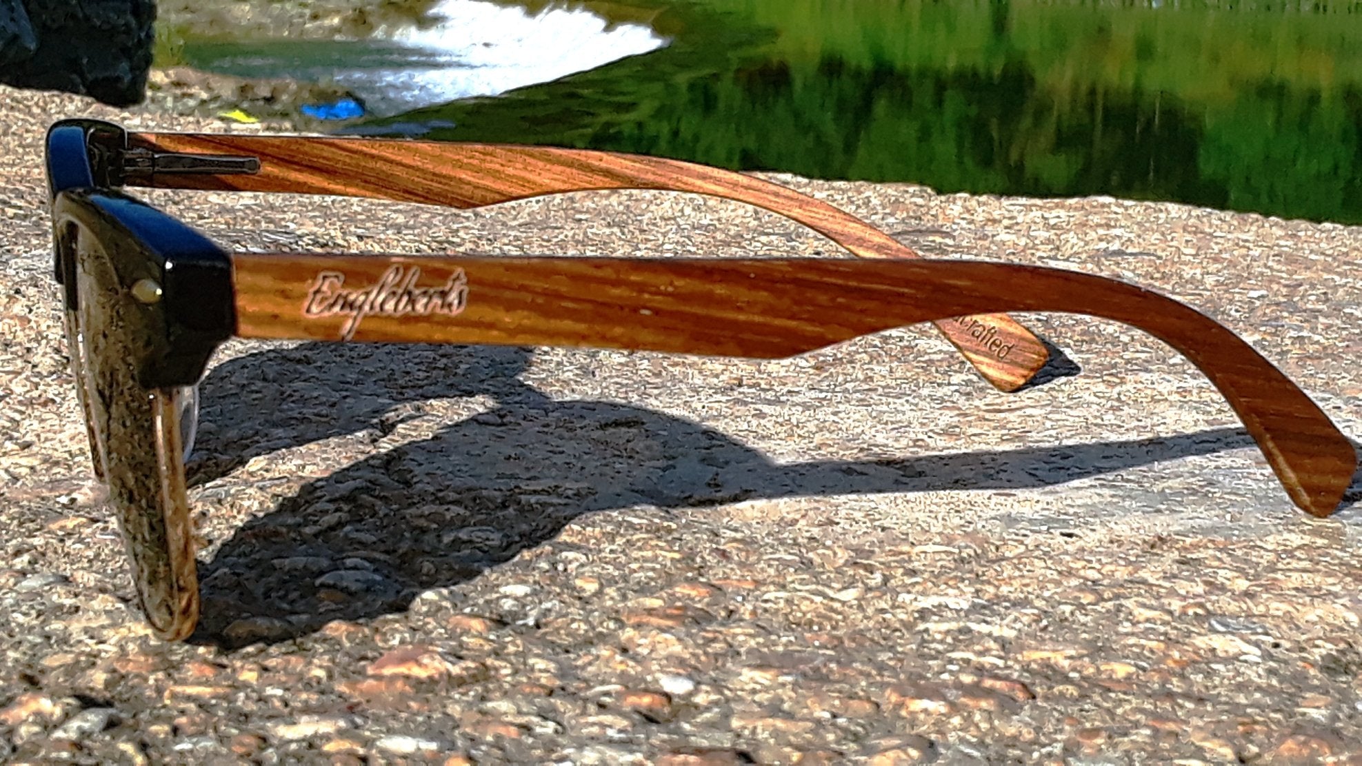 Wooden sunglasses with black frames rest on textured stone, greenery and water in the background.